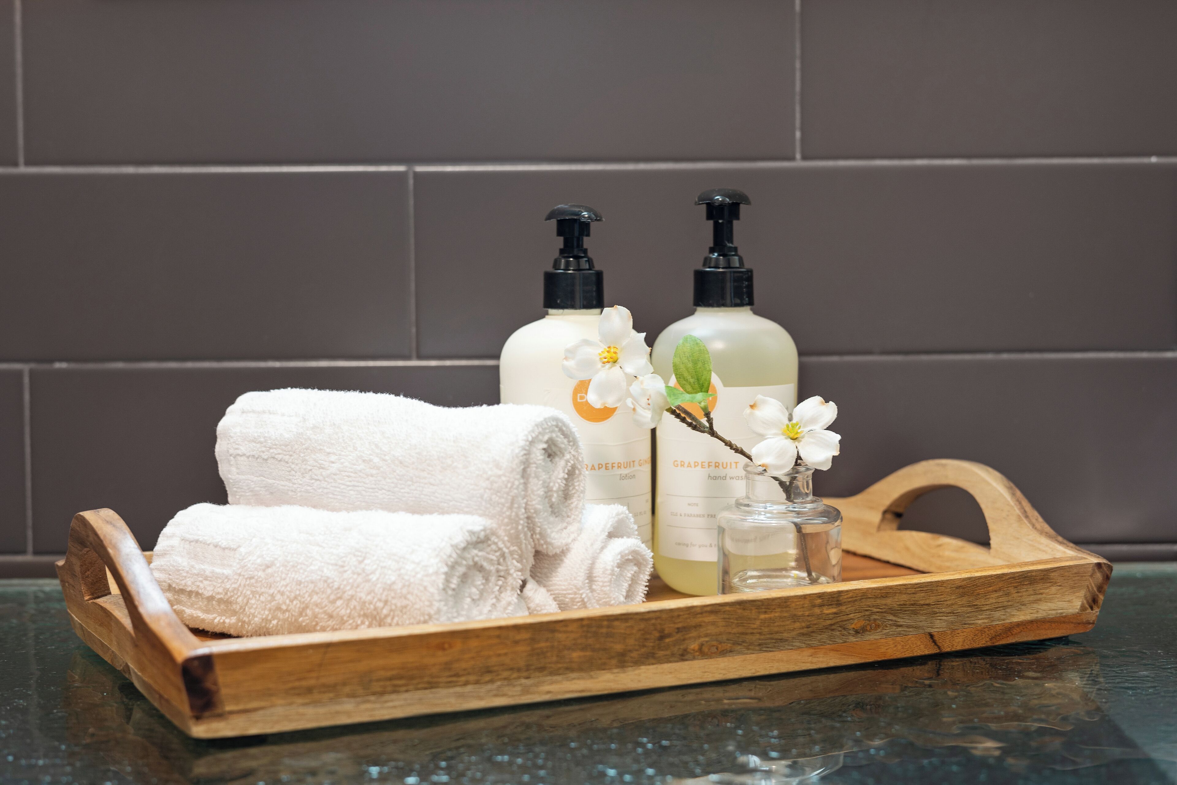 A wooden tray with two bottles of hand soap, a glass of liquid soap, and a rolled white towel on a reflective surface.