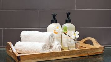 A wooden tray with two bottles of hand soap, a glass of liquid soap, and a rolled white towel on a reflective surface.