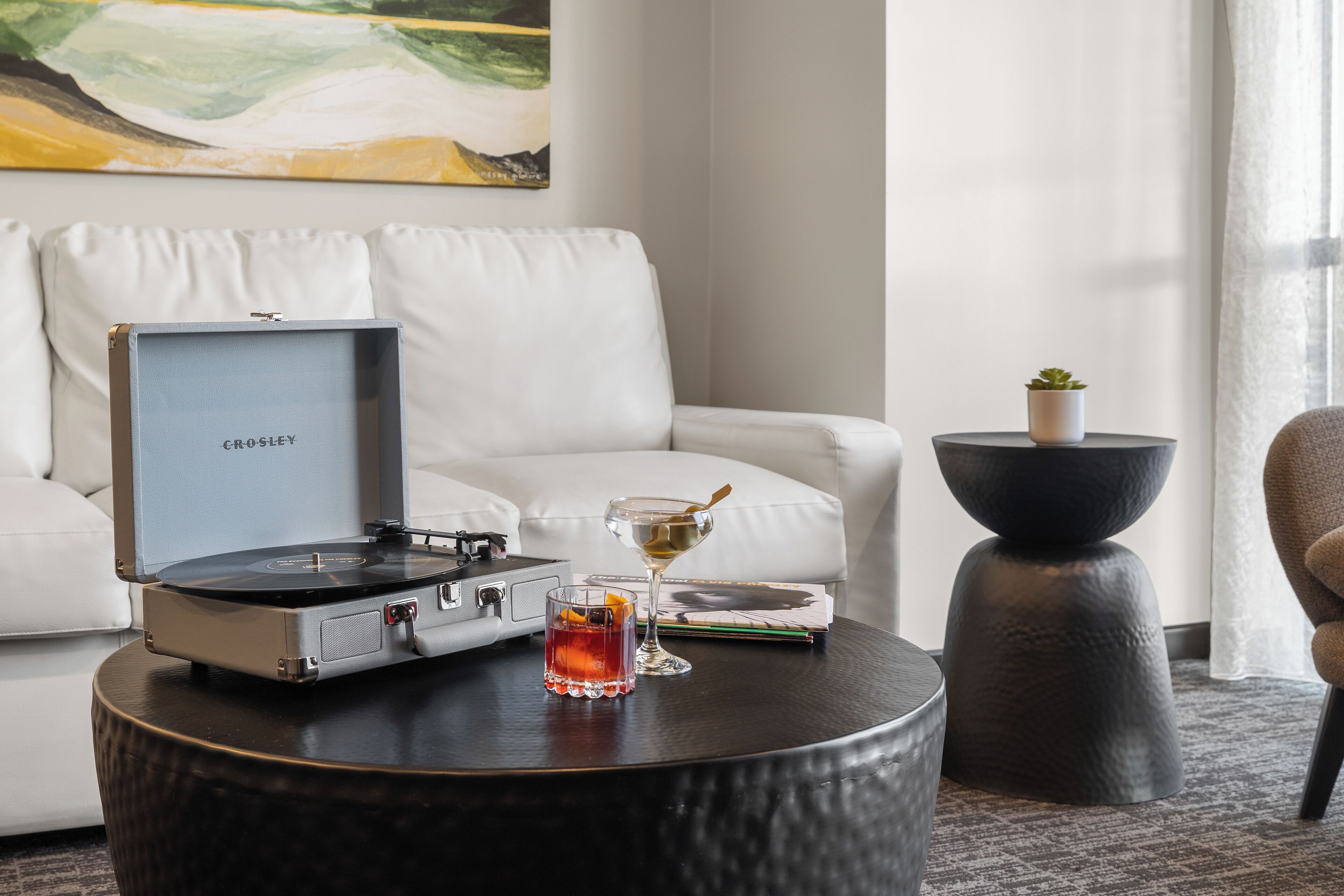 A modern living room with a white sofa, a round coffee table with a record player, a glass of whiskey, and a small potted plant.