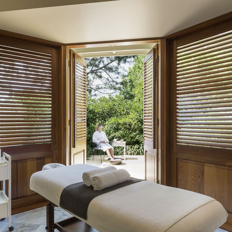 A spa room with a massage table, a small desk with a vase of flowers, and wooden blinds.