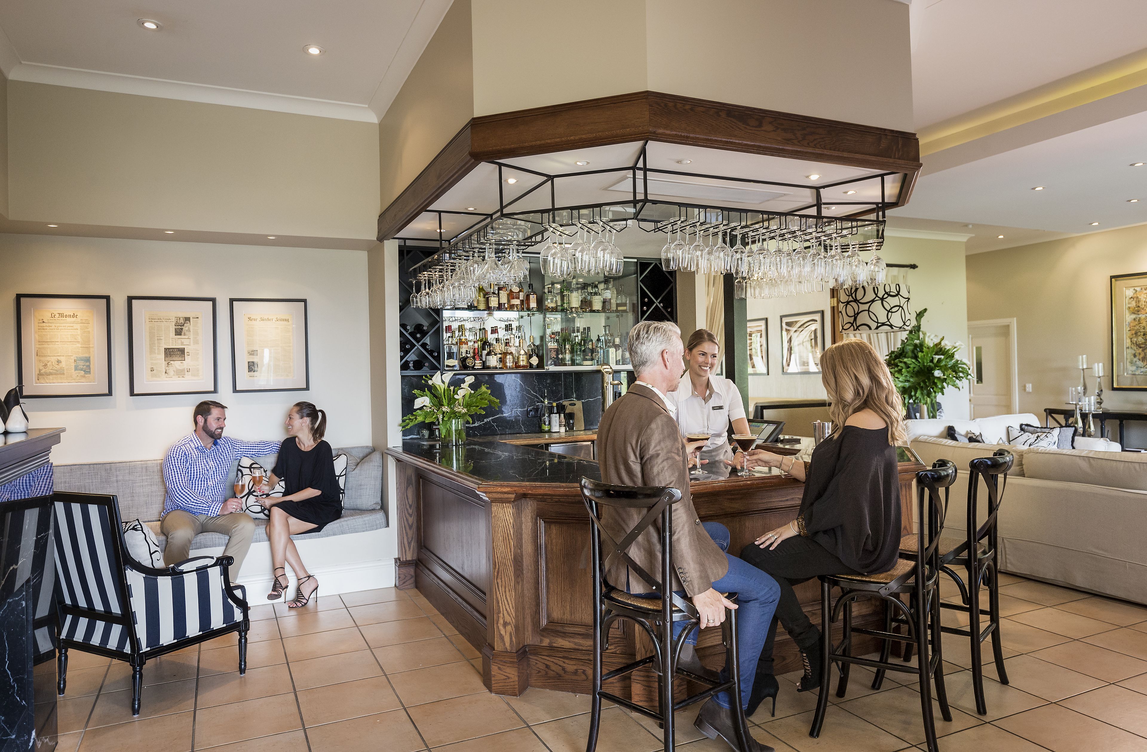A modern bar area with a wooden bar counter, hanging glassware, and a seating arrangement with bar stools and a sofa.