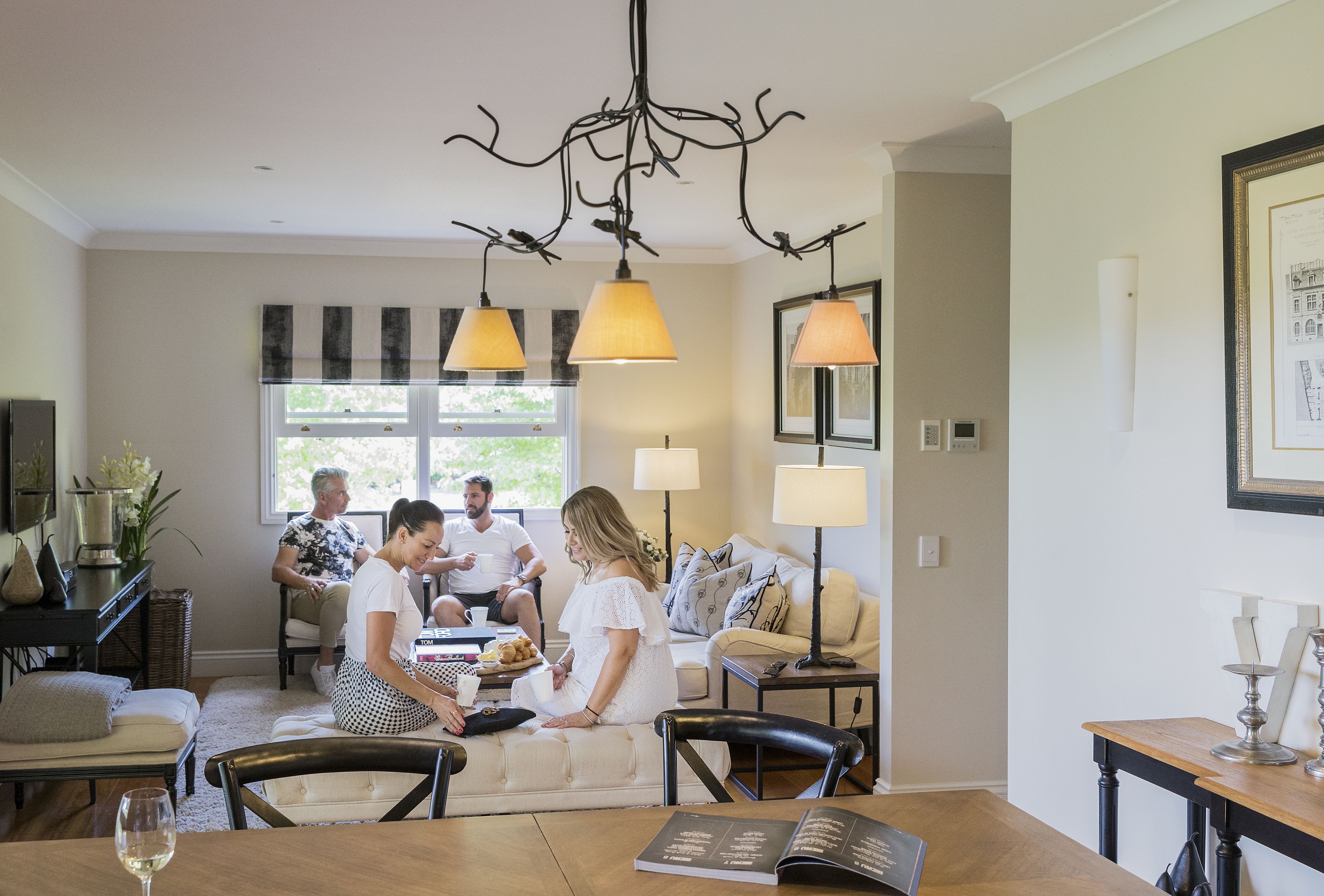 A dining area with a wooden table, chairs, and a chandelier. There is a sofa and a window with blinds in the background.