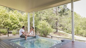 A couple sitting by a hot tub on a wooden deck with a view of trees and a fence.