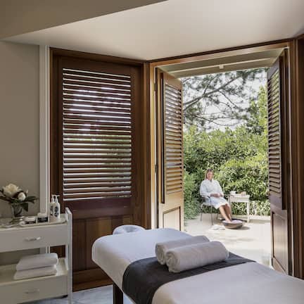 A spa room with a massage table, a small table with a vase and bottles, and a view of a person sitting outside.