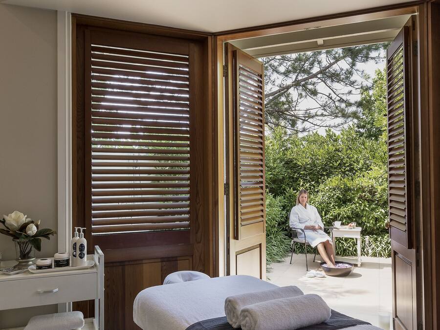 A spa room with a massage table, a small table with a vase and bottles, and a view of a person sitting outside.
