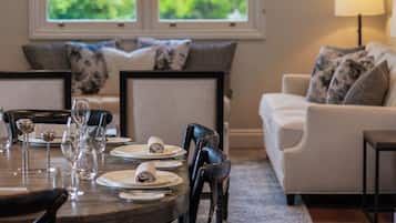 Un coin repas avec une table en bois dressée pour quatre personnes, entourée de chaises noires. Deux fenêtres offrent une vue sur la verdure à l’extérieur. Un canapé blanc avec des coussins à motifs est adossé au mur, et une lampe sur pied se trouve à droite.