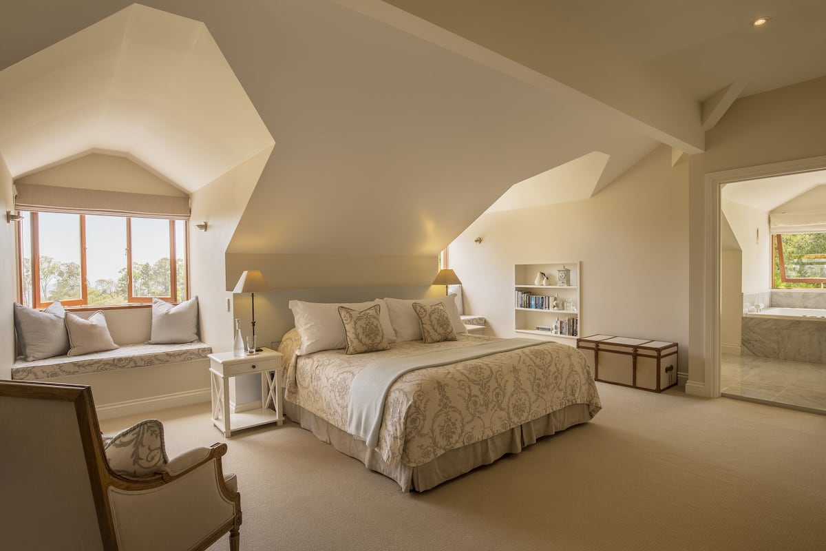 Une chambre avec un grand lit, un coin fenêtre aménagé en siège, une table de chevet, une commode, une bibliothèque et une salle de bain visible à travers une porte ouverte.
