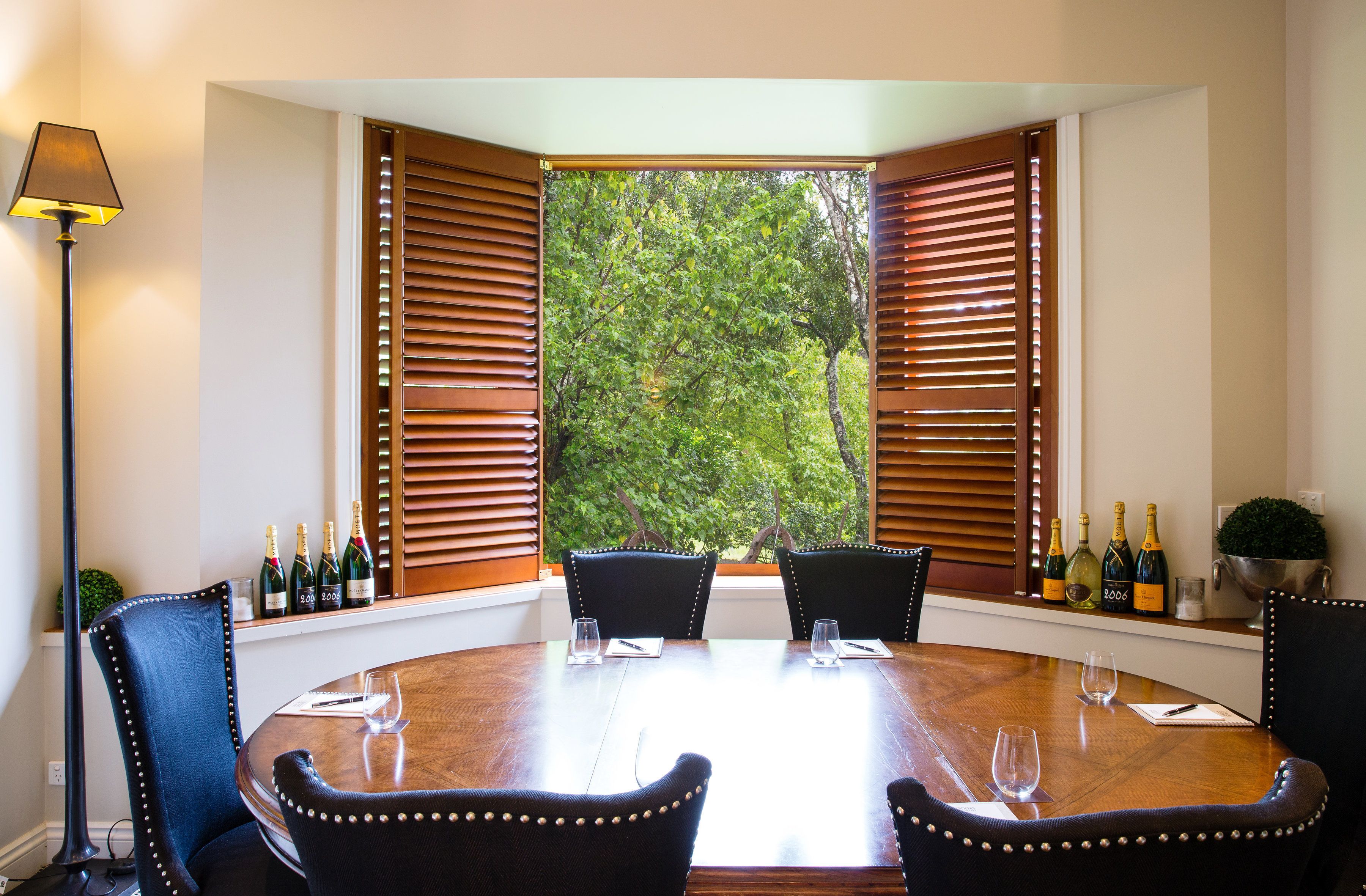 A dining area with a round table, four chairs, and a view through wooden shutters to a green outdoor area.
