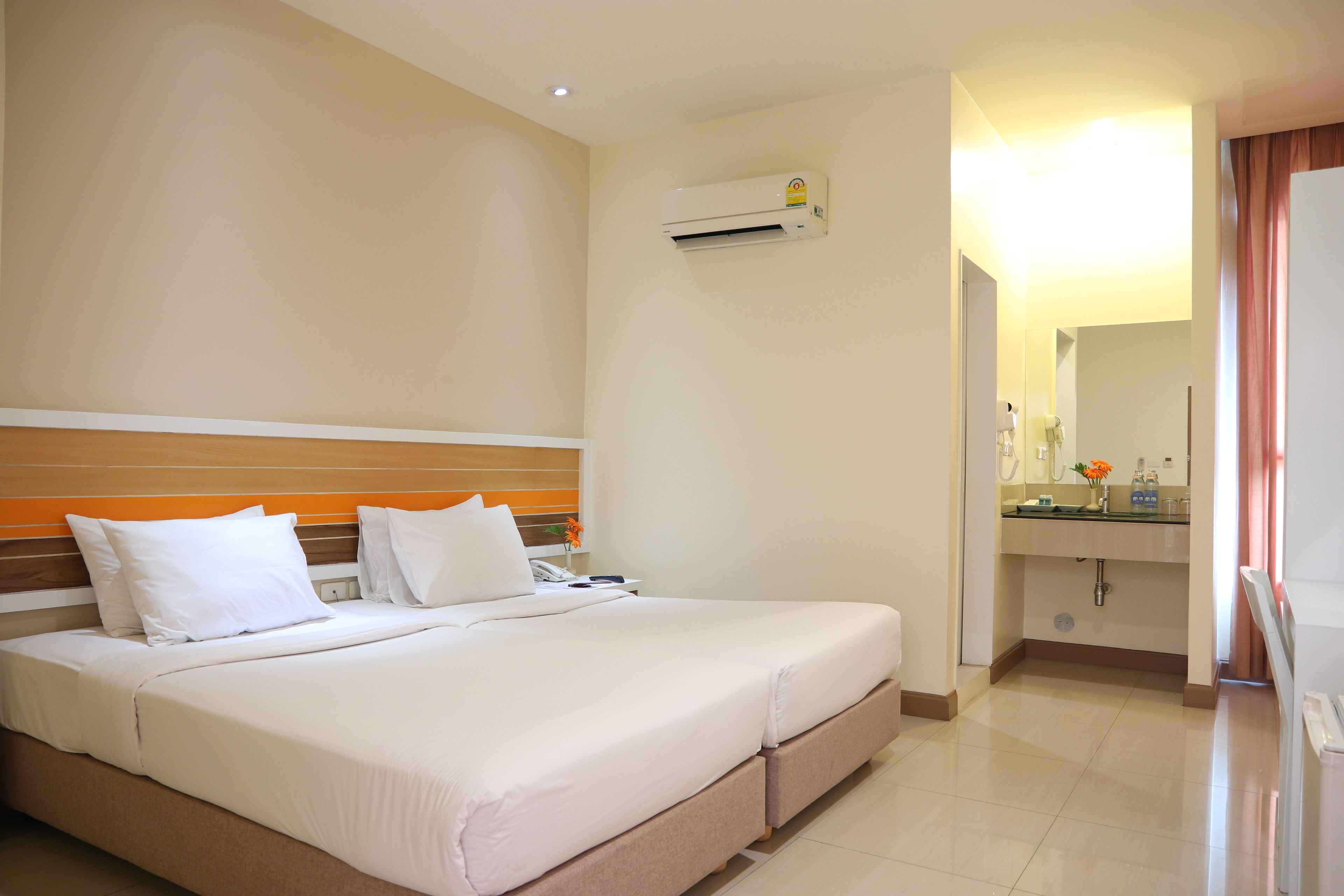 A hotel room with a large bed, a bedside table, an air conditioning unit, a bathroom with a sink and mirror, and a door leading to another room.