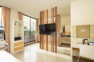 A modern hotel room with a flat-screen TV mounted on a wooden partition, a balcony with a view of the city, and a kitchenette area.