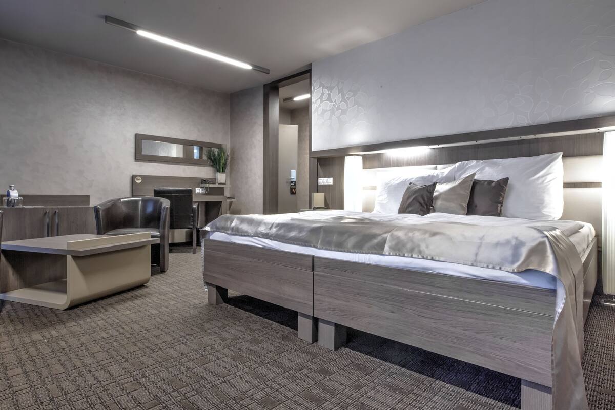 A modern hotel room with a large bed, a desk, a chair, and a small seating area.