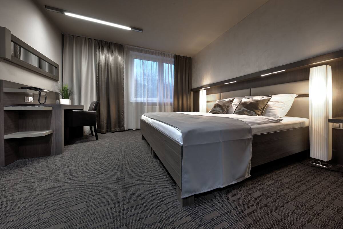 A modern hotel room with a large bed, a desk, and a chair. There is a plant on the desk and a window with curtains.