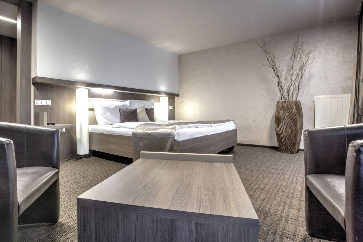 A modern hotel room with a large bed, two armchairs, a small table, and a vase with branches.