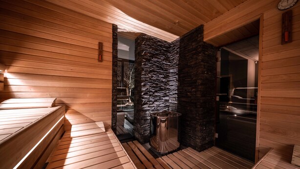 Ein Saunaraum mit Holzbänken, einer Steinwand und einer Glastür zum Außenbereich.