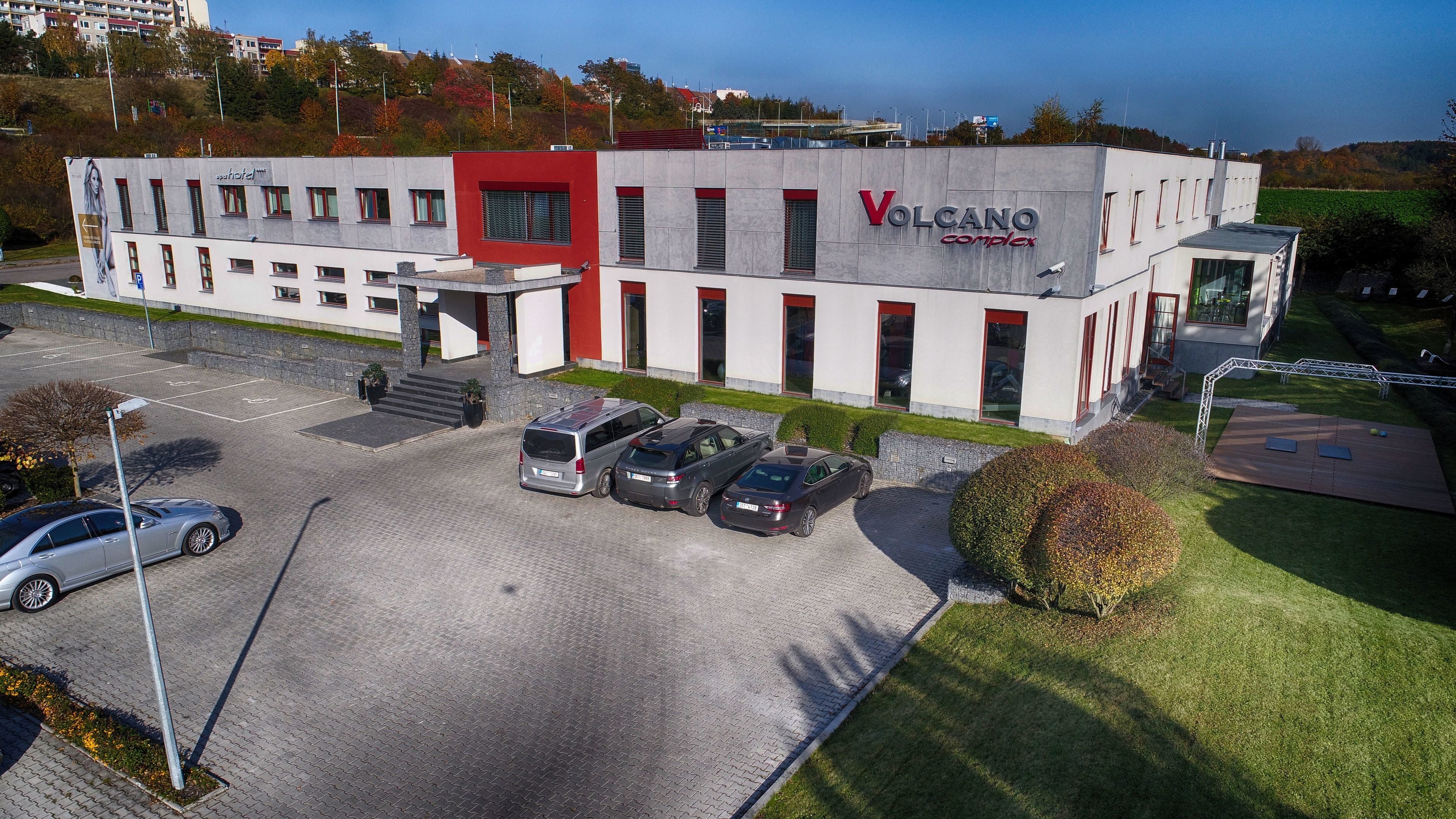 A building with a red and white facade, labeled "Volcano Complex," featuring a parking lot with several cars and a small outdoor area with a wooden deck.