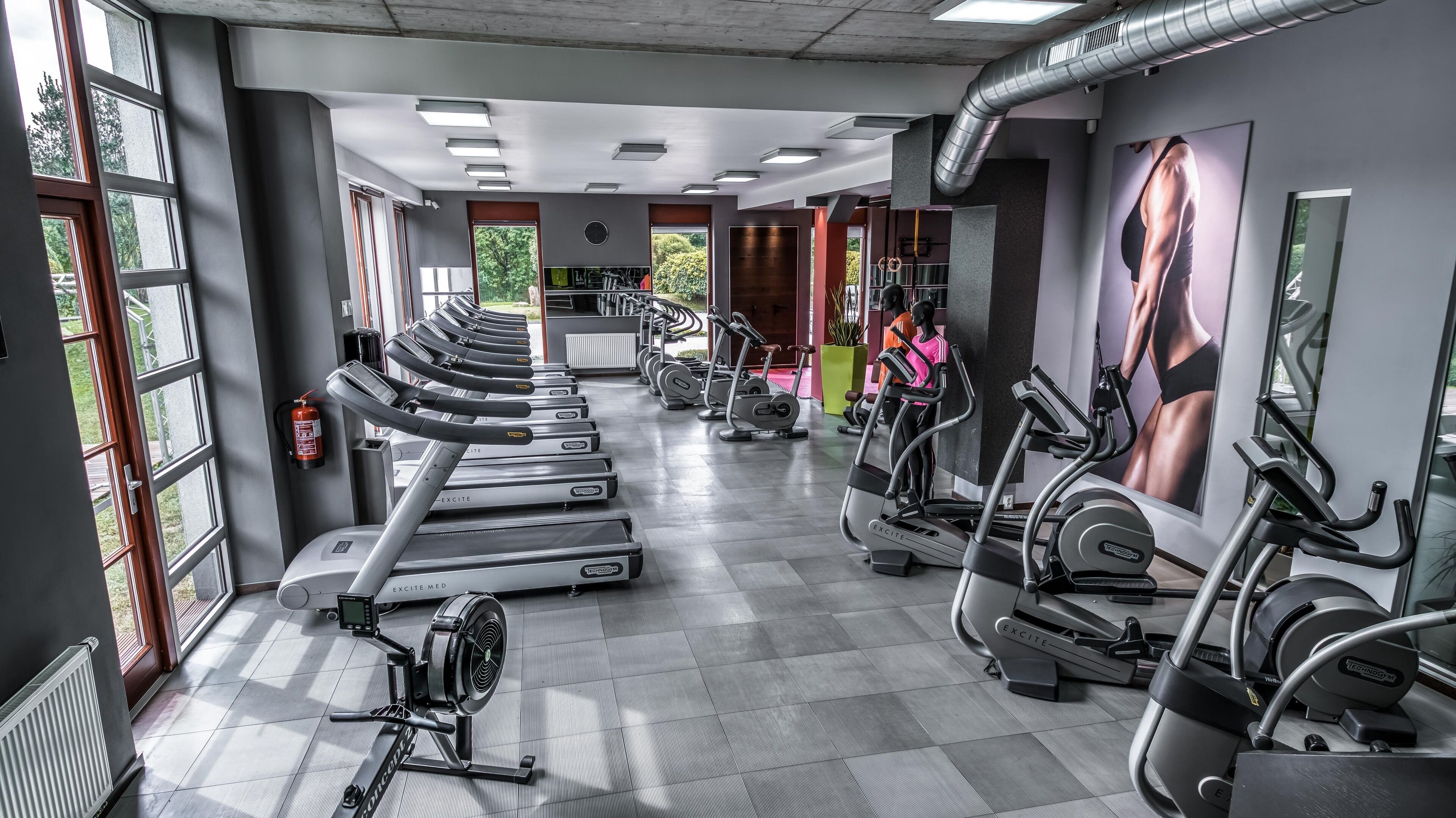 A gym with multiple treadmills and elliptical machines, large windows, and a view of trees outside.