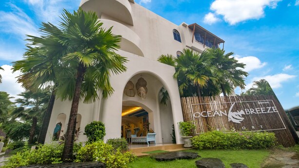 A resort with a white facade, arched entrance, palm trees, and a wooden fence with the resort's name. - Ocean Breeze Resort Khao Lak (Takua Pa)
