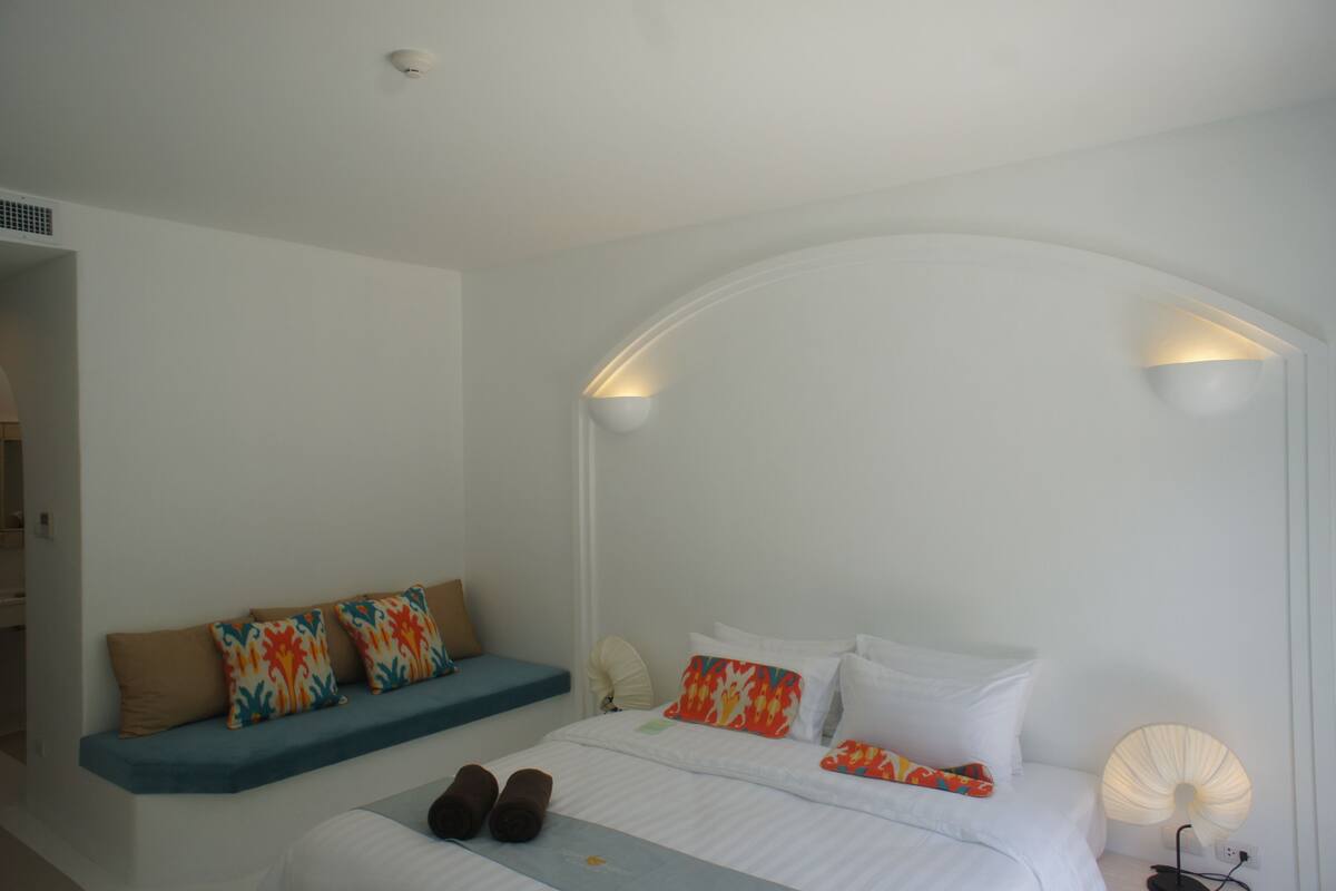 Een slaapkamer met een bed, kussens, een bankje en een wandlamp.