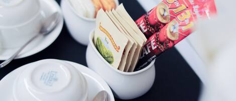 A table with white teacups and saucers, a bowl of tea bags, and a packet of instant coffee.