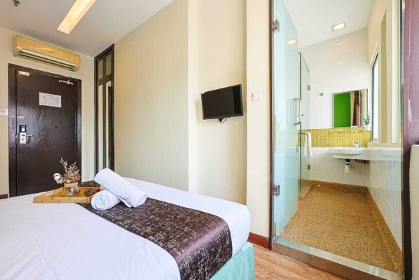 Habitación de hotel con cama, televisor, puerta y baño visible a través de un panel de vidrio.