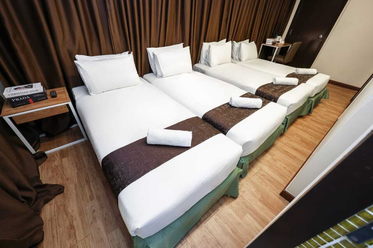 A hotel room with three beds, each with white linens and brown headboards. There is a small table with a book and remote control, and a curtain in the background.