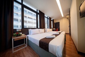 Habitación de hotel con dos camas, un escritorio, una silla, una mesita con una planta y vistas a edificios a través de amplios ventanales.