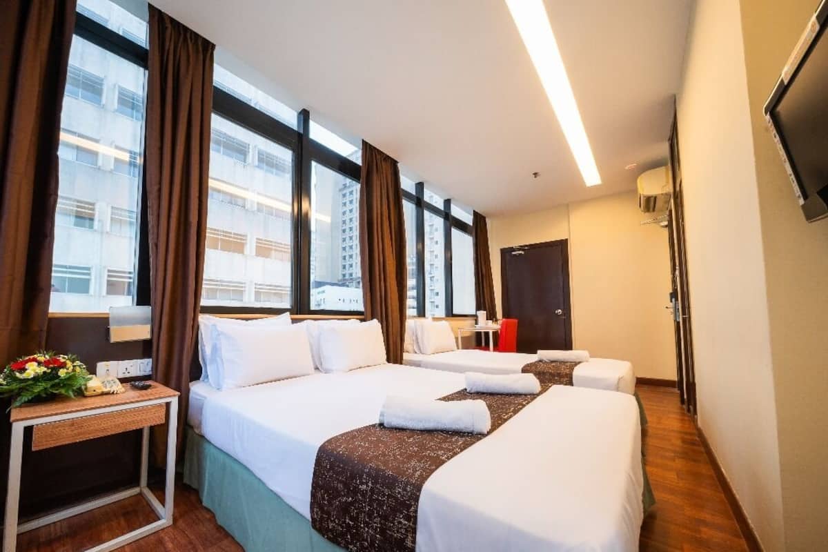 Une chambre d’hôtel avec trois lits, une table de chevet avec une lampe, un téléviseur fixé au mur et de grandes fenêtres avec des rideaux.