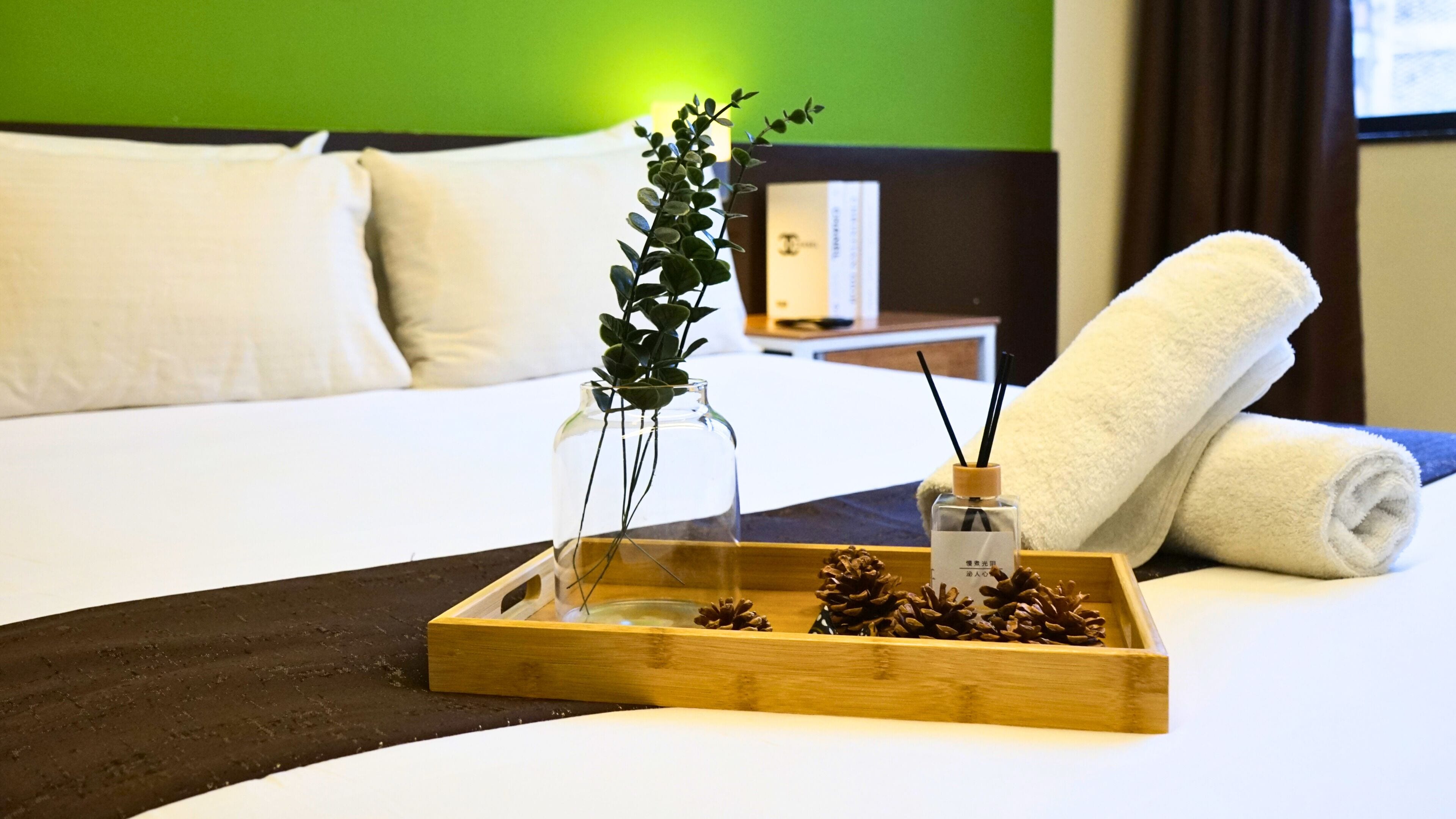 A neatly made bed with white linens, a wooden tray with pinecones and a small vase with greenery, and a white towel rolled up beside the bed.