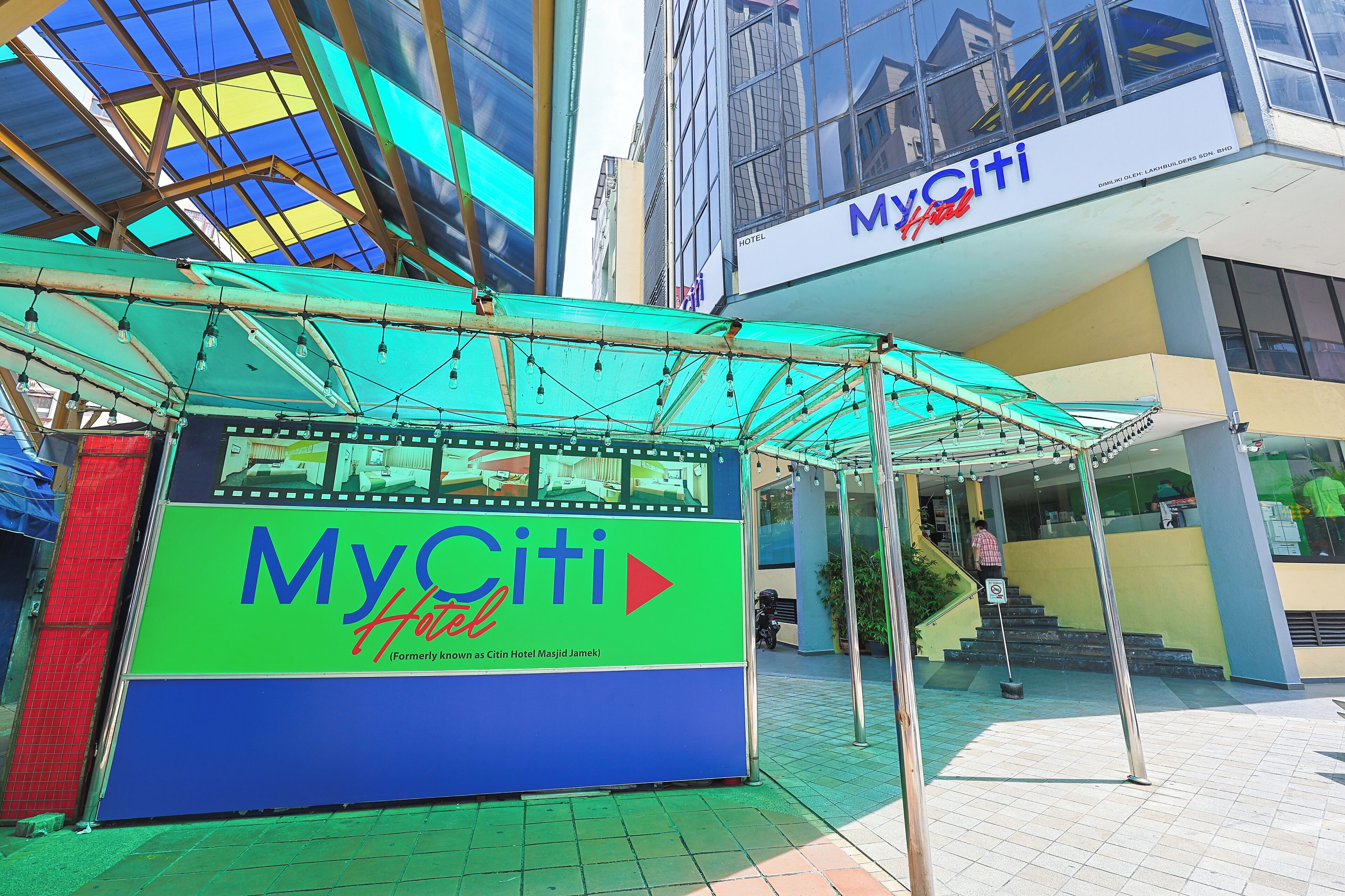Een bord van MyCiti Hotel met een blauw- en groen kleurenschema, gelegen in een commercieel gebied met een glazen gebouw op de achtergrond.