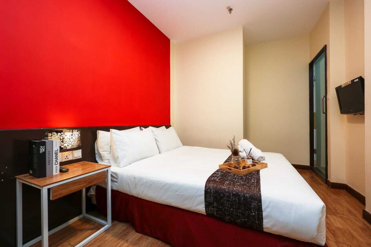 A hotel room with a large bed, a bedside table with a book and a remote control, a red accent wall, and a television mounted on the wall.
