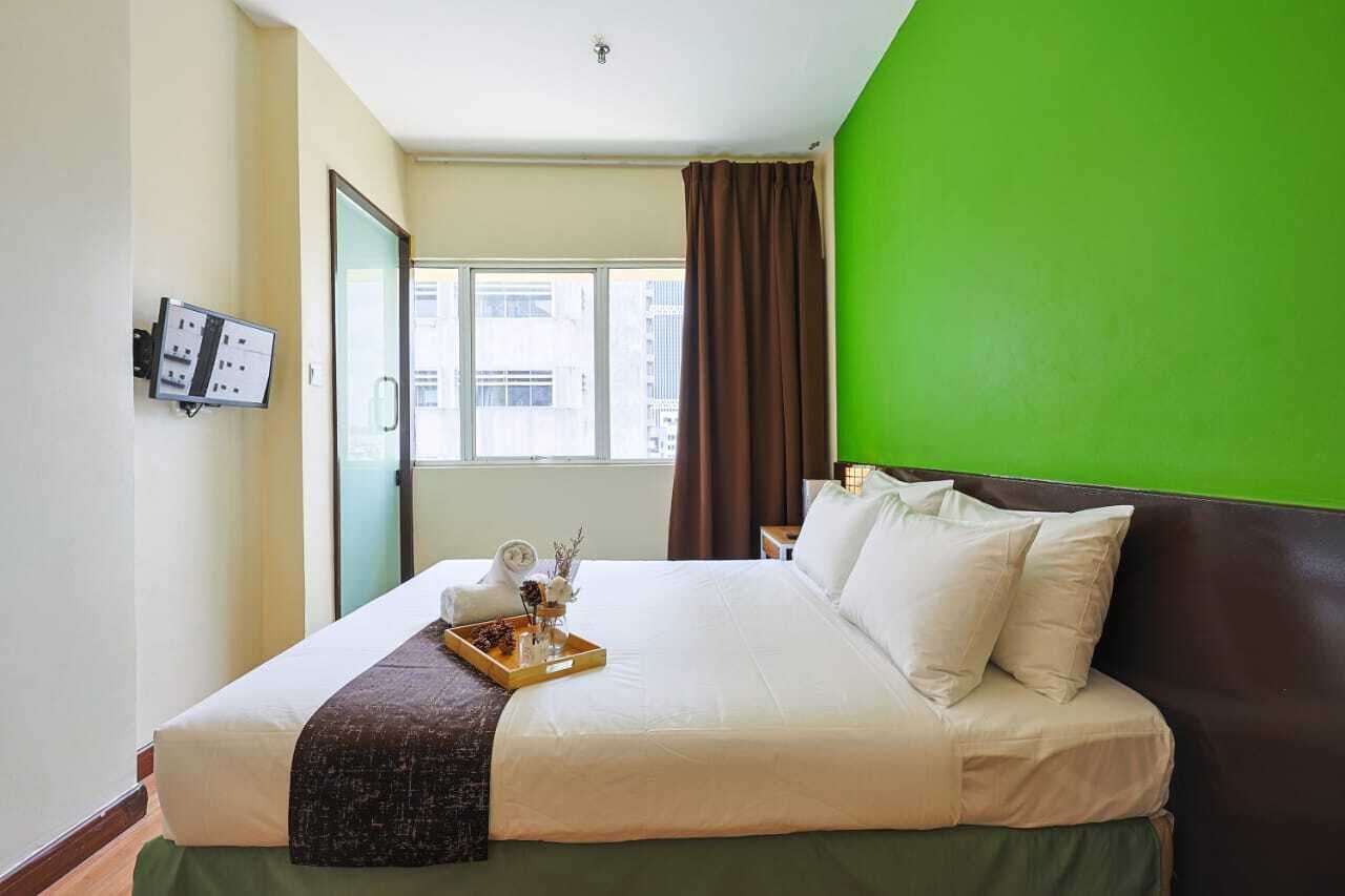 A hotel room with a bed, a green wall, a window with curtains, and a wall-mounted control panel.