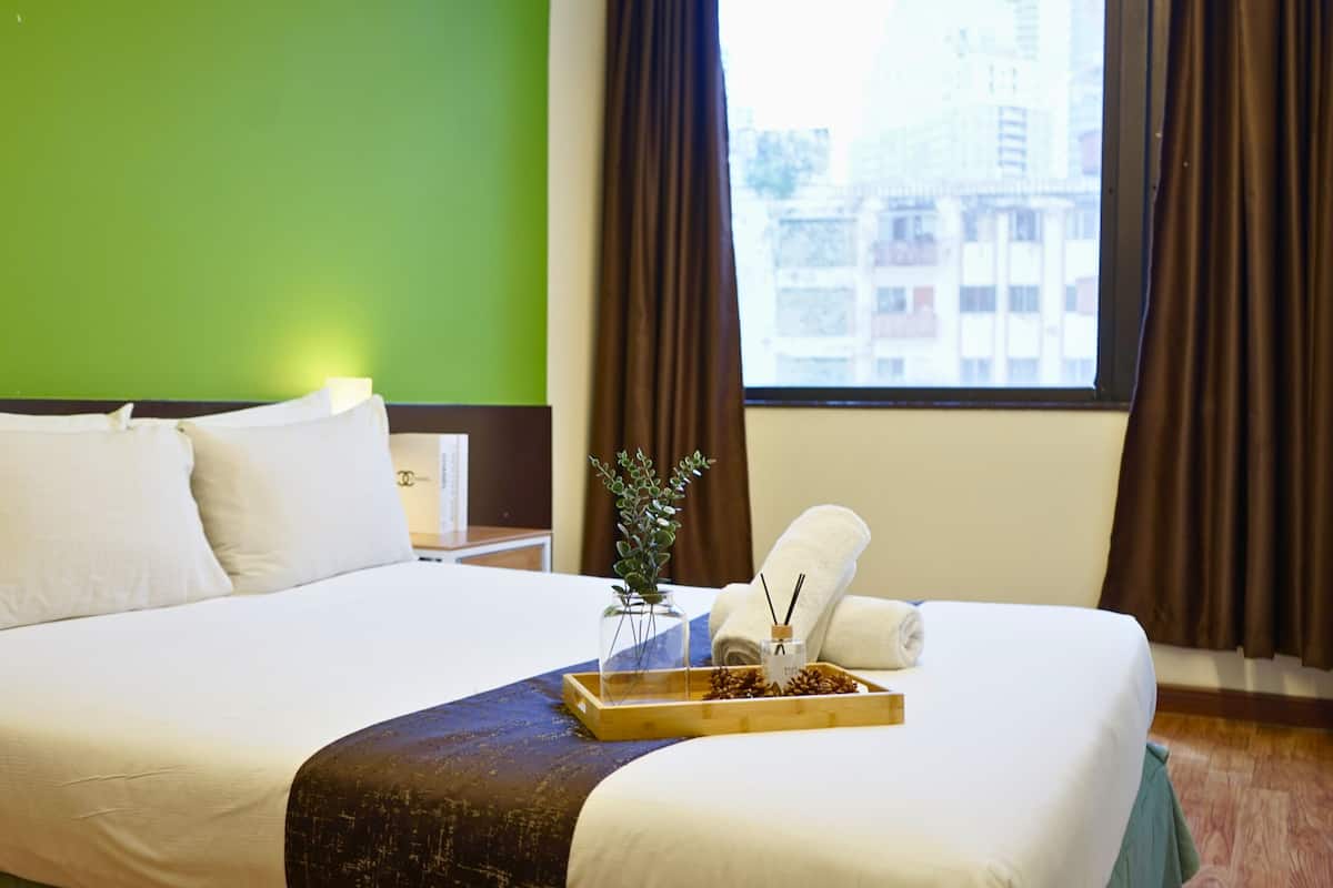 Une chambre d’hôtel avec un grand lit, des murs verts, une fenêtre avec des rideaux et un plateau contenant une serviette et une petite plante.