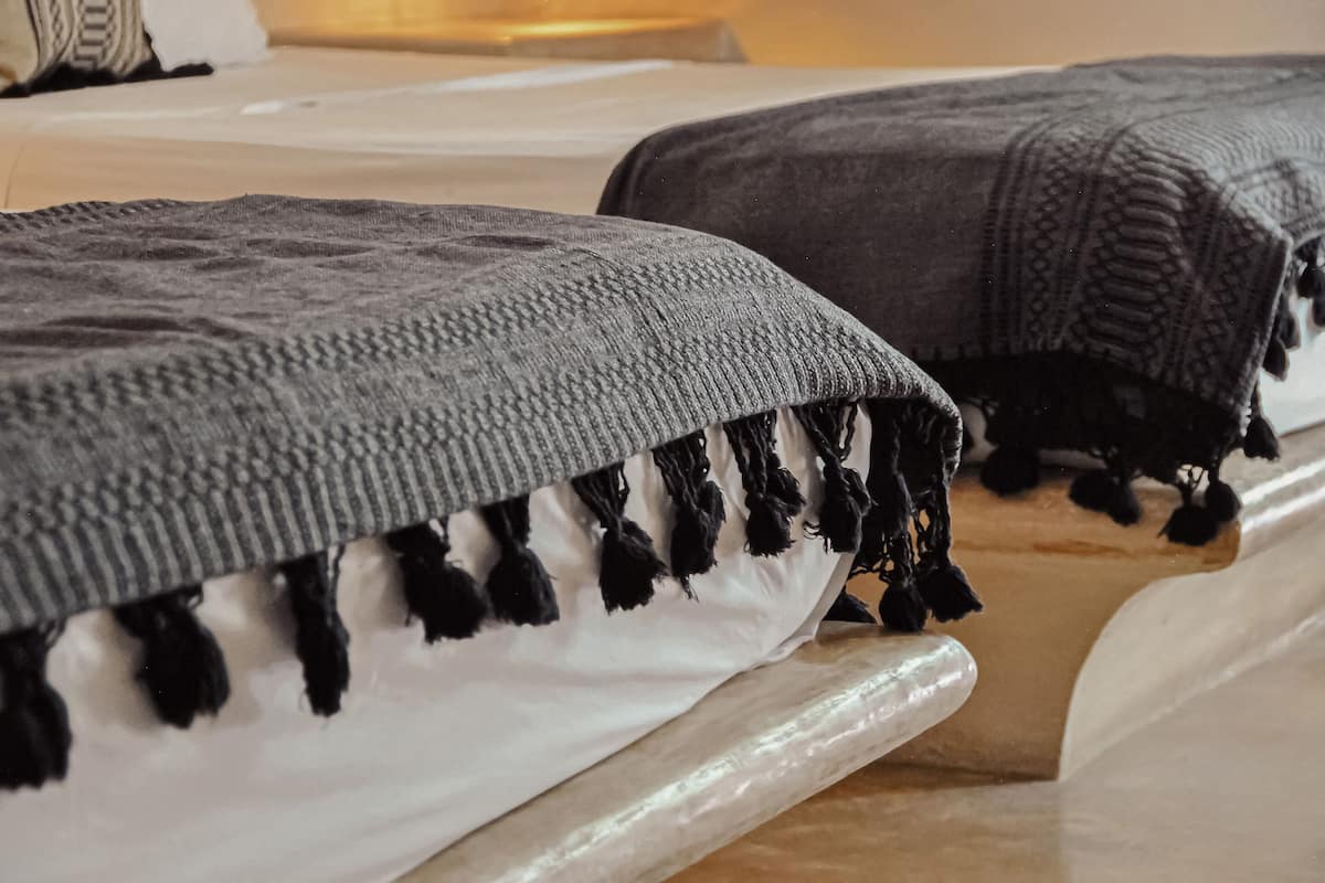A bed with a grey knitted blanket and black tassels, a white pillow, and a wooden headboard.