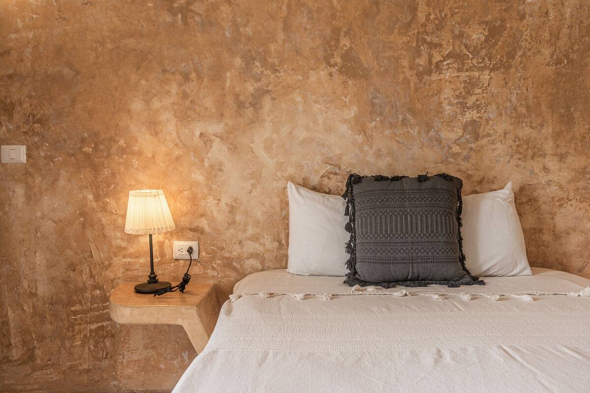 A bedroom with a textured wall, a bedside table with a lamp, and a bed with white bedding and patterned pillows.