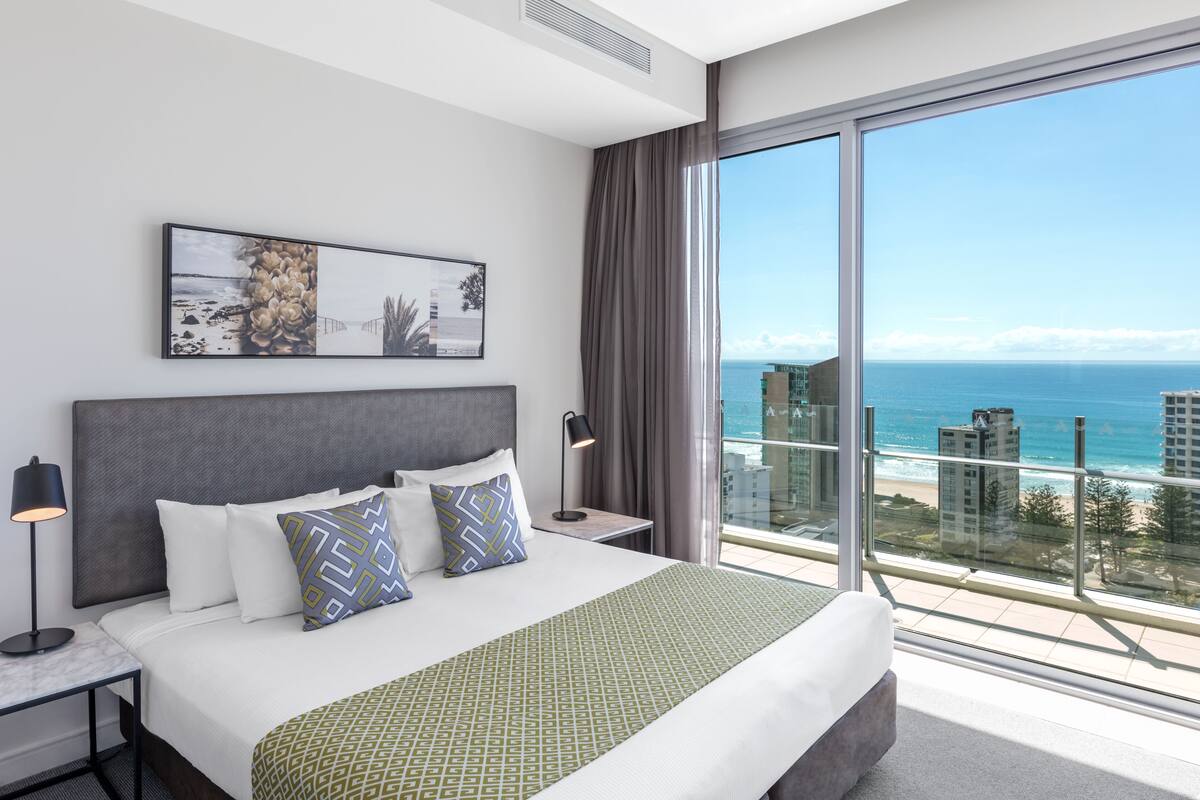 A hotel room with a large bed, a nightstand, a lamp, a framed picture above the bed, and a view of the ocean through a large window.