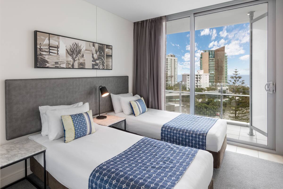 A hotel room with two beds, a large window with a city view, a framed artwork on the wall, and a nightstand with a lamp.