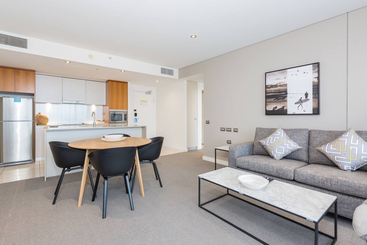 A modern living room with a grey sofa, black chairs, a marble coffee table, and a kitchen area with wooden cabinets and a stainless steel refrigerator.