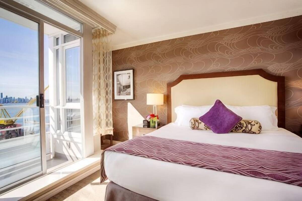 A hotel room with a large bed, a nightstand, a lamp, and a window with a view of a cityscape.
