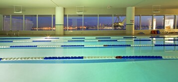 Una piscina interior con carriles delimitados, amplias ventanas con vistas a la ciudad y una grúa visible en el exterior.
