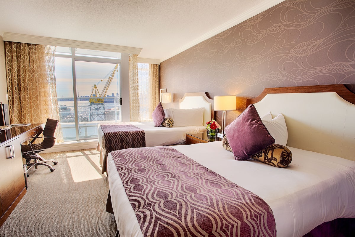A hotel room with two beds, a desk, and a view of a cityscape.
