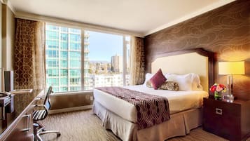 A hotel room with a large bed, a desk, and a chair. There is a window with a view of buildings outside.