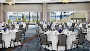 A banquet hall with round tables set for dining, large windows offering a view of a bridge and cityscape, and a modern chandelier. - Pinnacle Hotel at the Pier (North Vancouver)