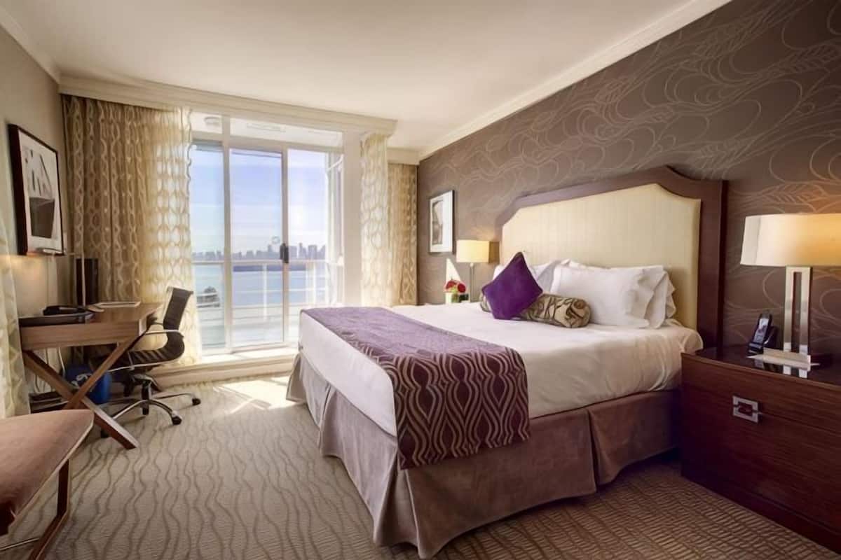 A hotel room with a large bed, a desk, a chair, a lamp, and a view of the city.