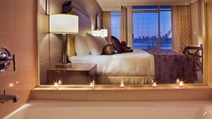 A hotel room with a large bed, bedside lamps, a nightstand, a view of the city through large windows, and a bathtub with candles. - Pinnacle Hotel at the Pier (North Vancouver)