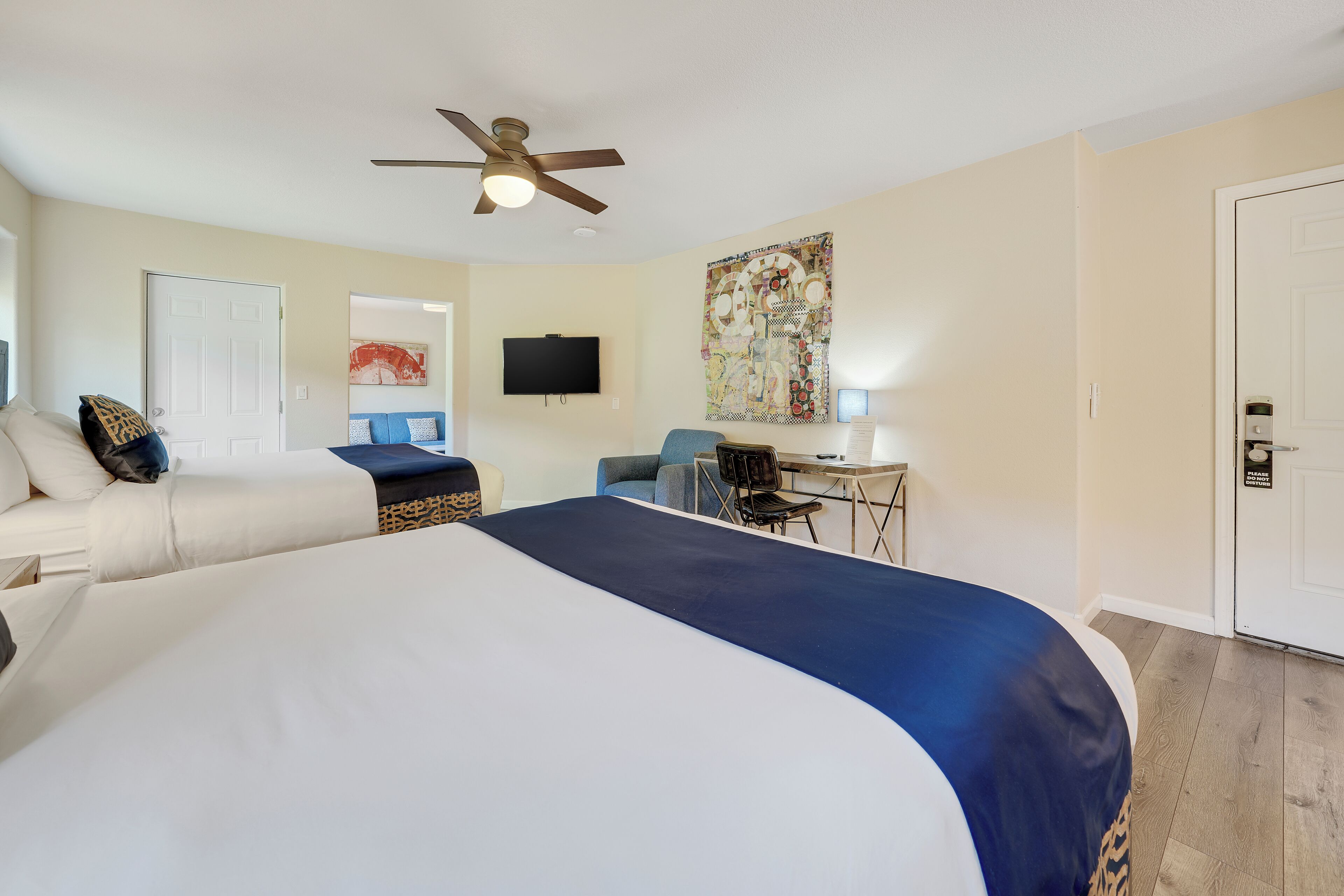 A hotel room with a large bed, a desk, a chair, a ceiling fan, a television, and a door with a 'Please Do Not Disturb' sign.