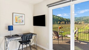 Una habitación con escritorio, silla y televisor montado en la pared. Hay una puerta corrediza de vidrio que da a una terraza exterior con vistas a una zona boscosa.