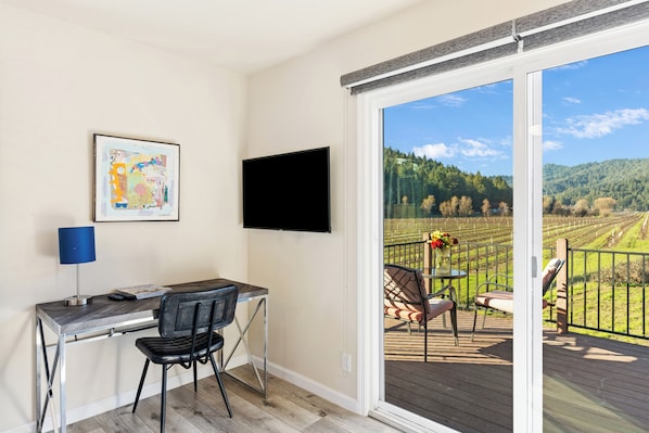 A room with a desk, chair, and a TV mounted on the wall. There is a sliding glass door leading to an outdoor deck with a view of a forested area.