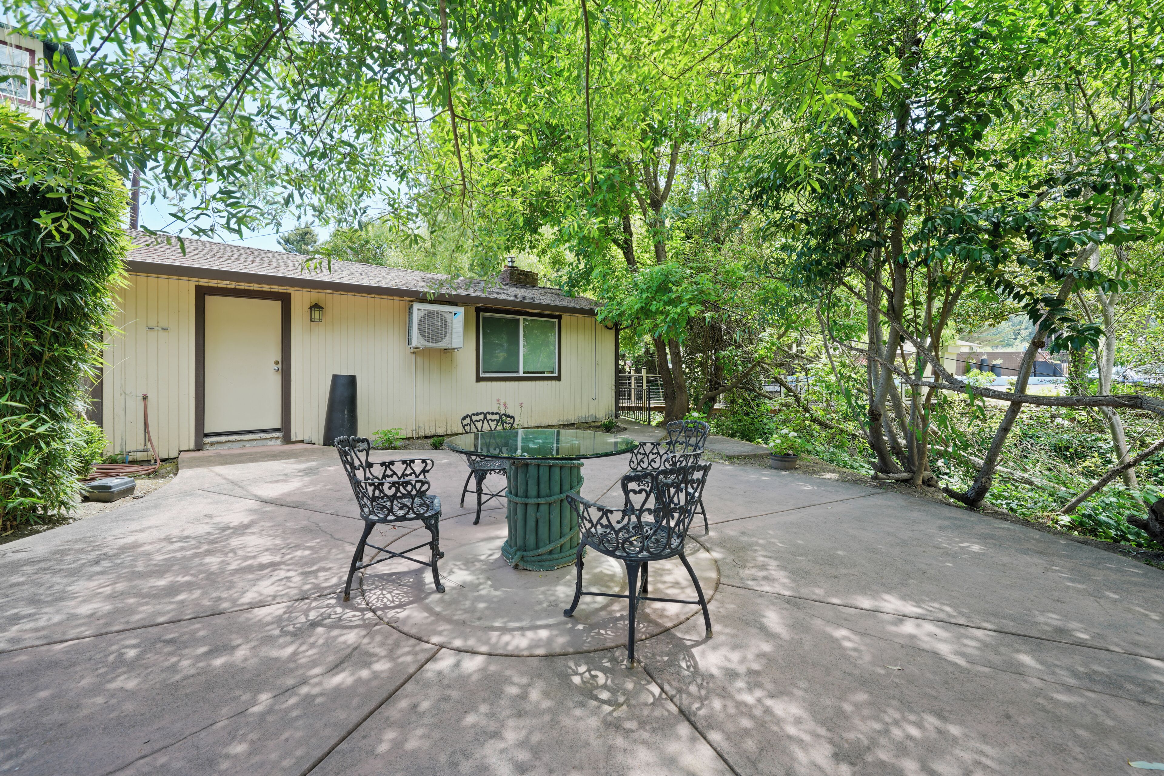 Une terrasse avec une table ronde et quatre chaises, un petit bâtiment doté d’une porte et d’un appareil de climatisation, le tout entouré d’arbres.
