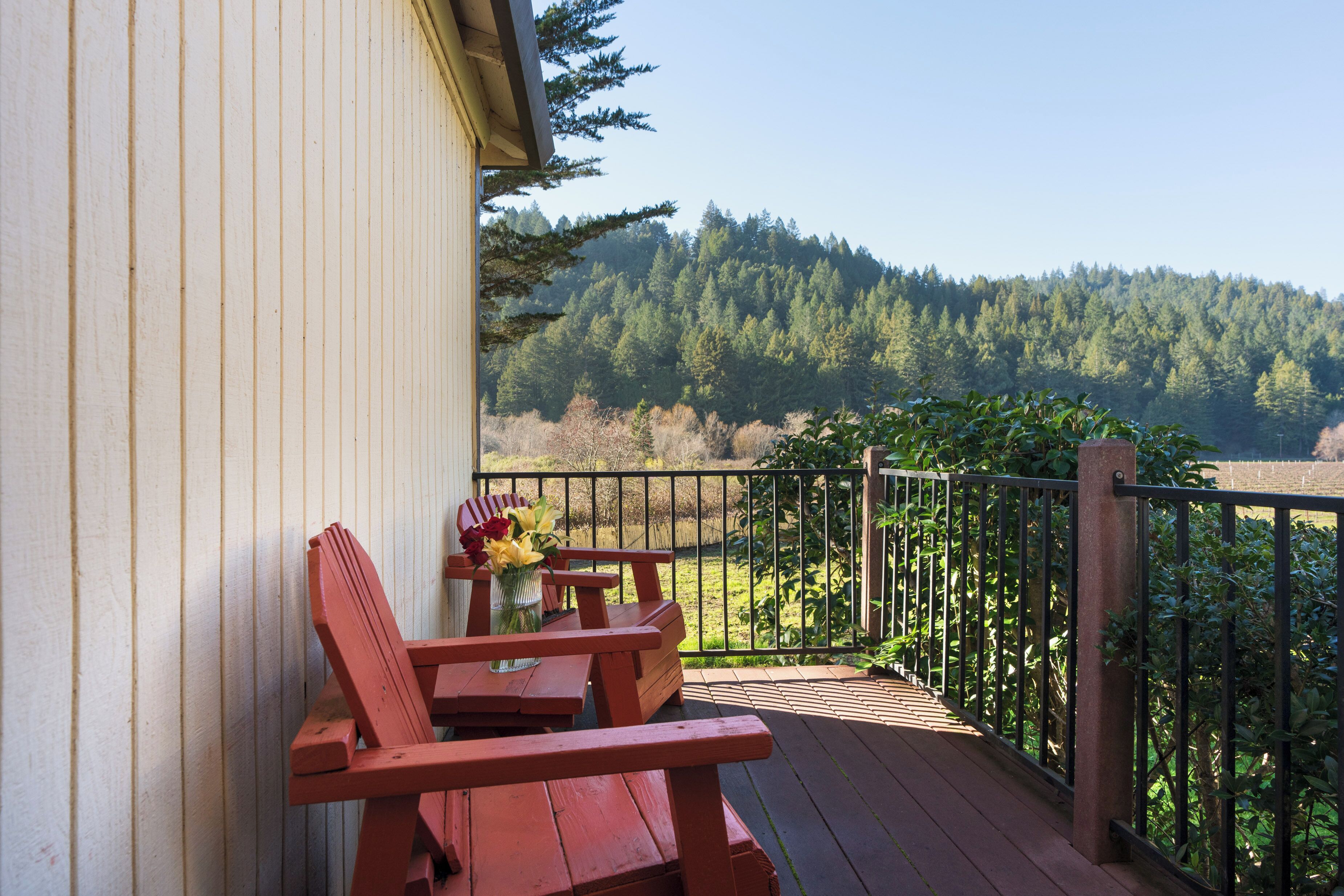 A wooden deck with red Adirondack chairs and a table, overlooking a forested hillside.