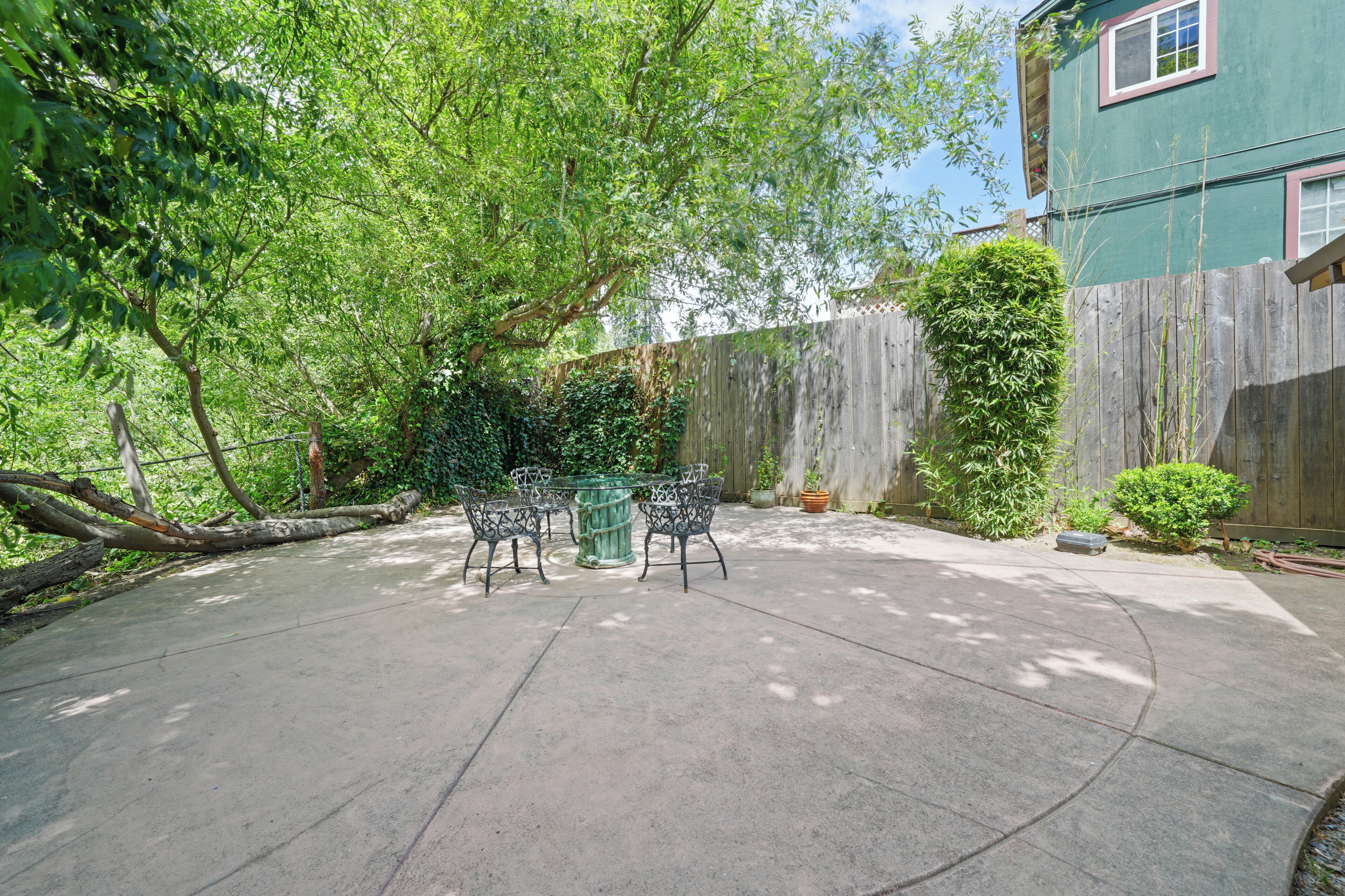 A concrete patio with a table and chairs, surrounded by a wooden fence and lush greenery.